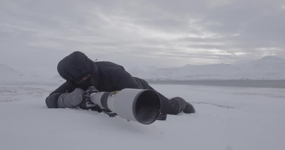 A photographer lies in the snow and adjusts his long zoom lens