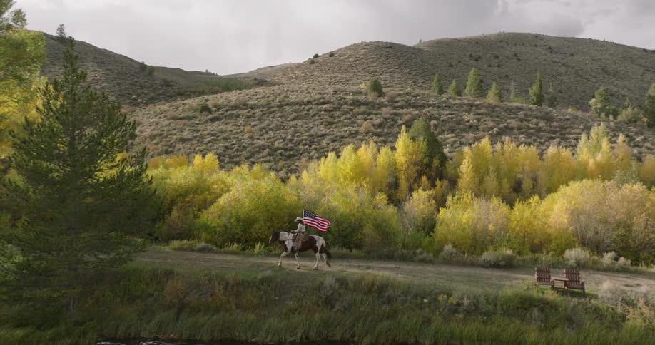 Person riding on a horse carries an american flag as they travel on a path at the base of the mountain