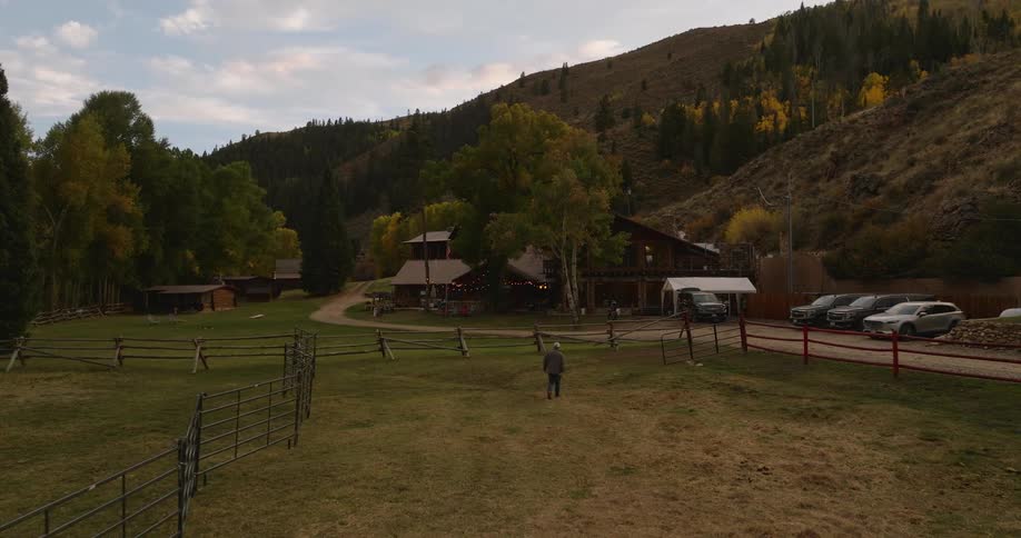 A person walks toward a rustic ranch house nestled among trees and hills with parked cars and fenced enclosures in the foreground