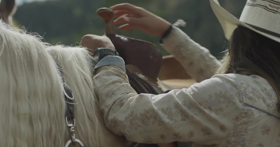 Woman in cowgirl hat adjusting leather belts on a white horse