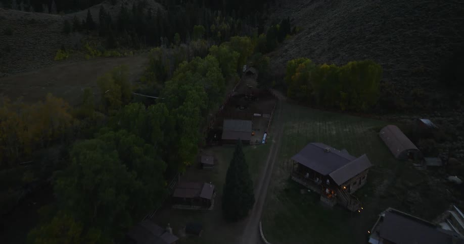 As dusk settles a drone offers an aerial view of a rustic ranch nestled in a valley with scattered cabins a winding dirt road and surrounding hills displaying hints of autumn colors