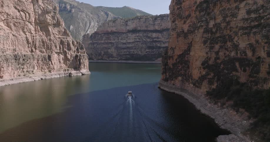 A powerboat creates a wake as it travels through a wide canyon with towering rock formations on either side