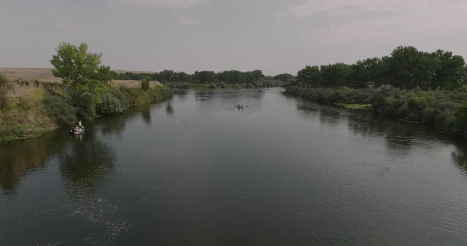 A small boat floats down a calm river surrounded by trees and fields