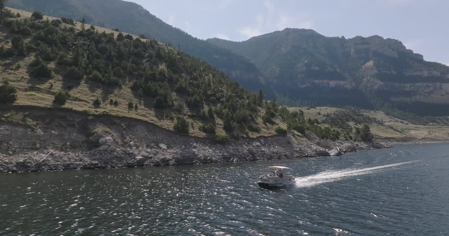 A pontoon boat moves across a body of water leaving a wake with a rocky tree covered hillside and mountains in the background under a partly cloudy sky