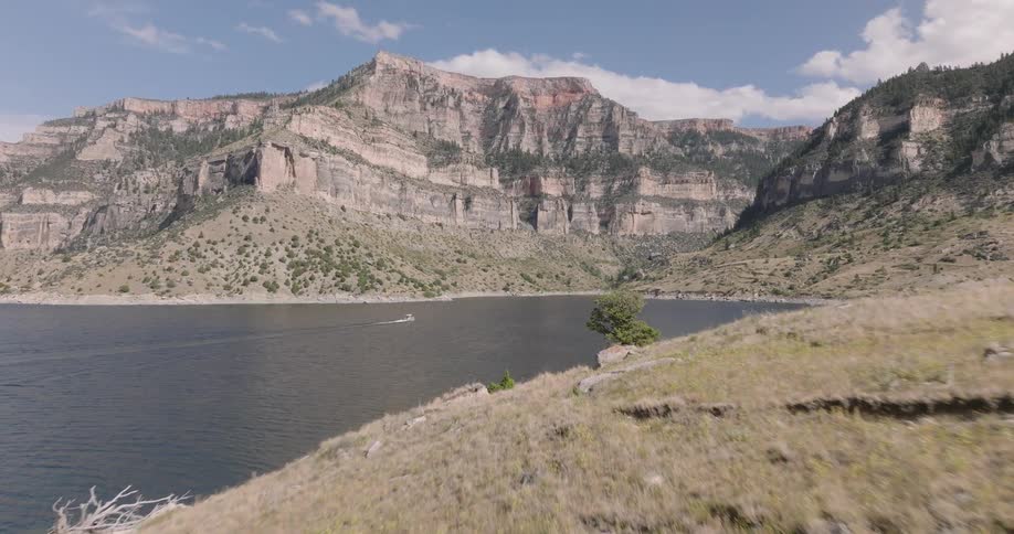 A boat moves across a wide lake surrounded by rocky cliffs and grassy slopes