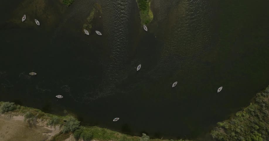 An aerial view shows several small white boats spread across a dark river with green vegetation along its banks and small islands in the water