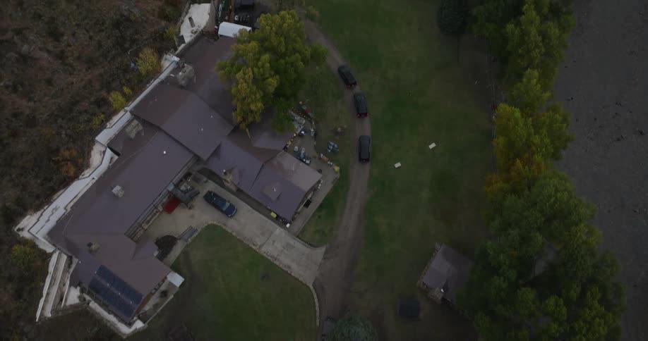 An aerial view shows a cluster of buildings with dark brown roofs surrounded by green grass and scattered trees with dirt paths traversing the property