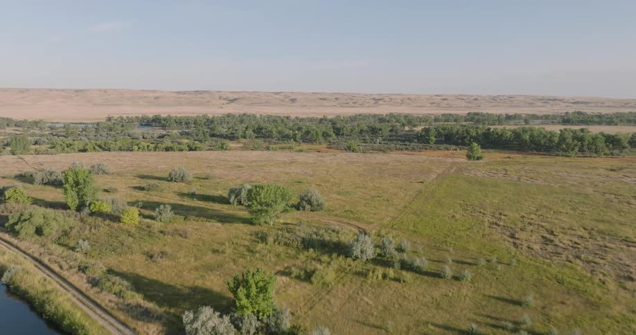 A forwardmoving aerial view glides over an irrigation canal and a grassy plain towards distant sandy hills on a hazy day