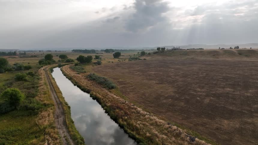 An aerial view shows a long canal reflecting the overcast sky as it flows through rural farmland and fields under dramatic sun rays breaking through the clouds
