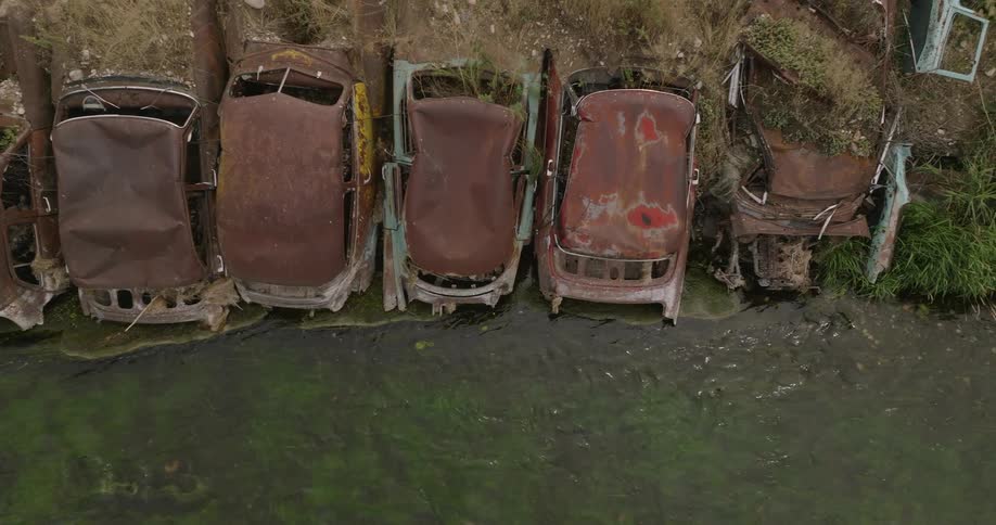 Rusty abandoned cars line the edge of a green grassy riverbank