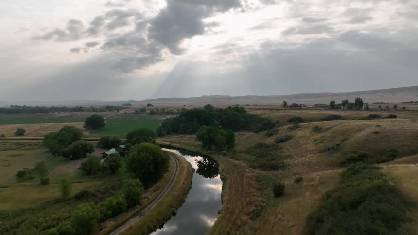 A winding river cuts through open fields under dramatic sun rays piercing the clouds