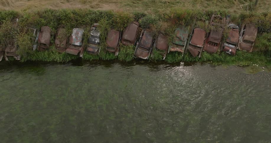 Aerial view of rusted abandoned cars lined up tightly along a grassy riverbank