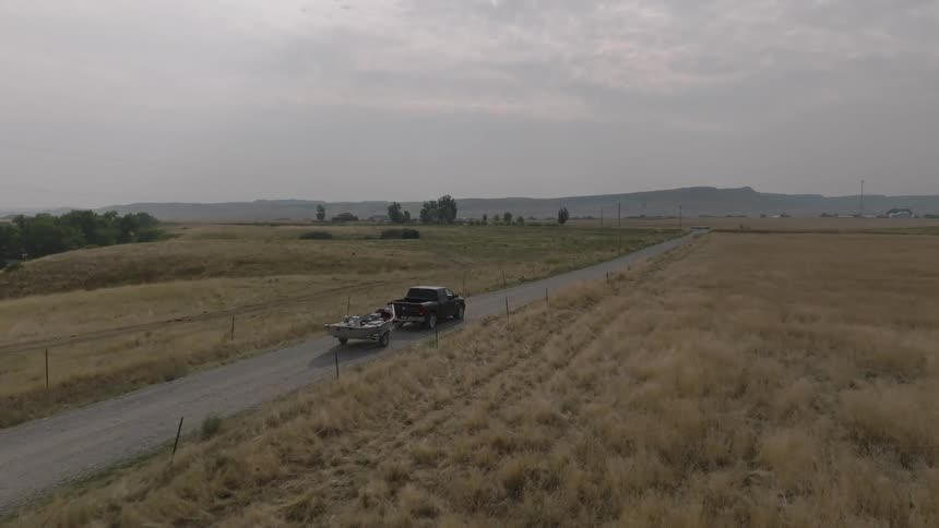A pickup truck is seen towing a boat down a rural dirt road that passes through dry fields under an overcast sky