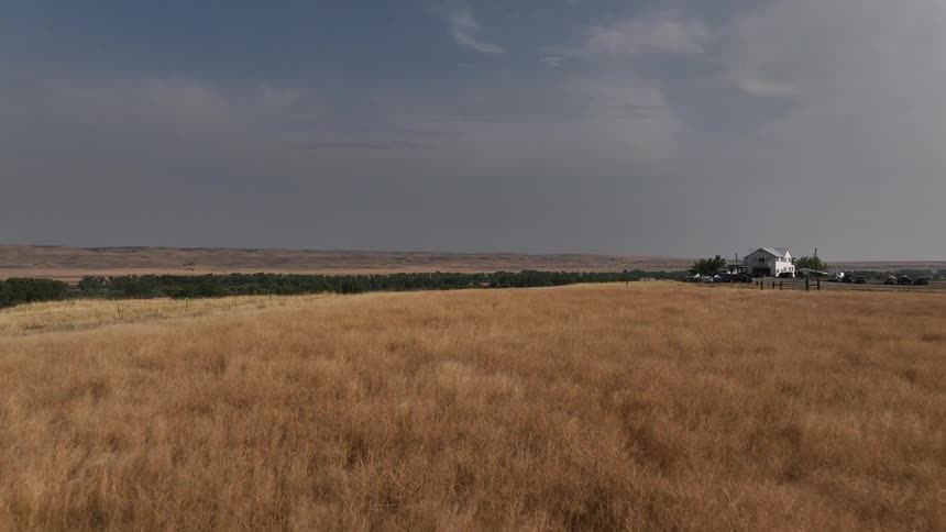 A dry grassy field stretches toward a river and a farmhouse under a cloudy sky