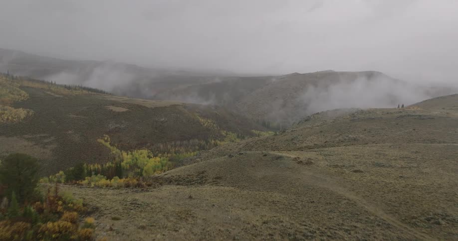 A drone flies over a rugged arid landscape dotted with patches of autumn colored trees as mist drifts through the distant mountain valleys under an overcast sky