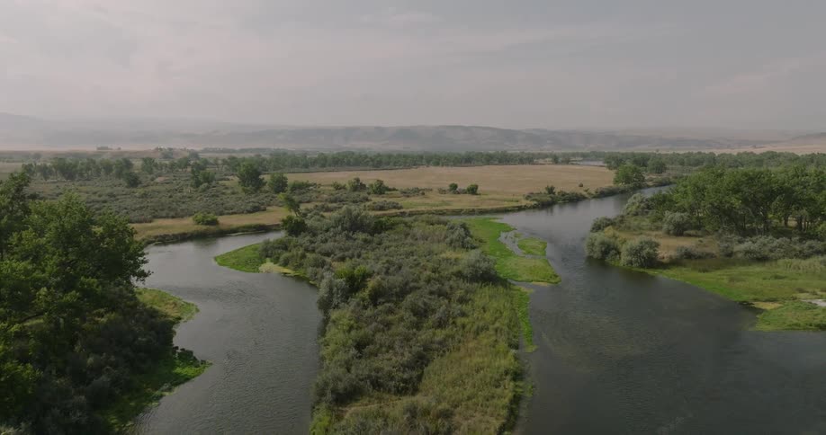 Aerial view of a winding river flowing through green fields and trees under a hazy sky