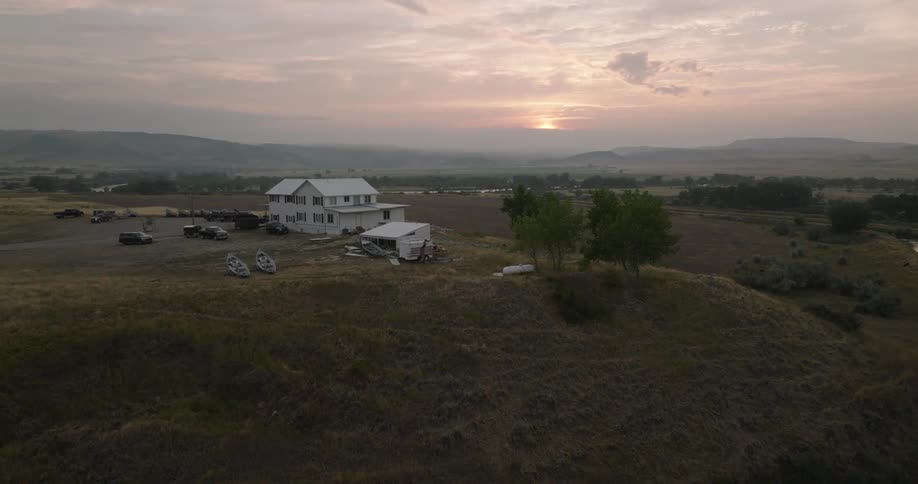An aerial shot shows a rural farm house situated on a hill with boats on the ground a river in the foreground and a sunrise illuminating the distant mountains