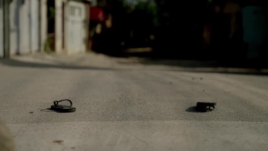 A worn out soccer ball rolls in the middle of an empty street flanked by a pair of sandals