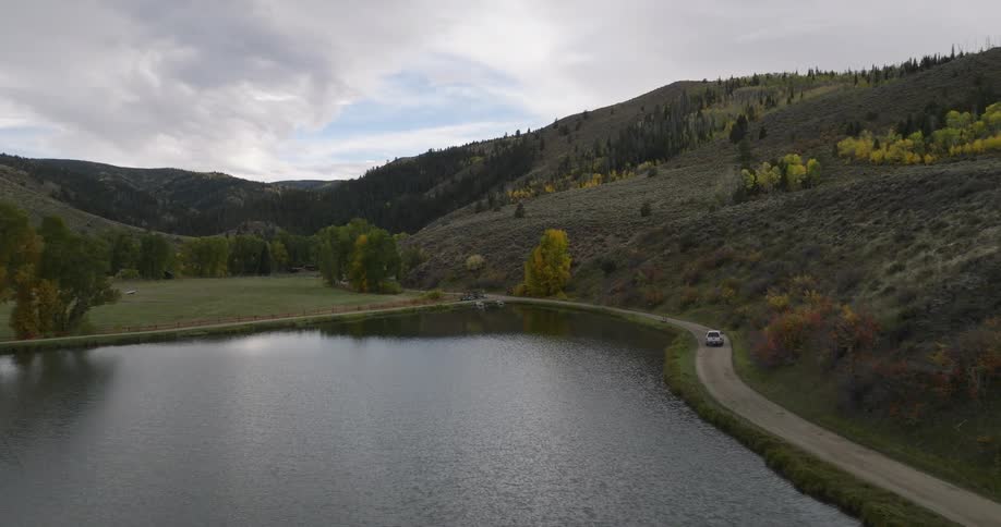 An aerial view shows a car driving along a dirt road next to a tranquil lake with hillsides covered in a mix of green and golden autumn trees under a cloudy sky