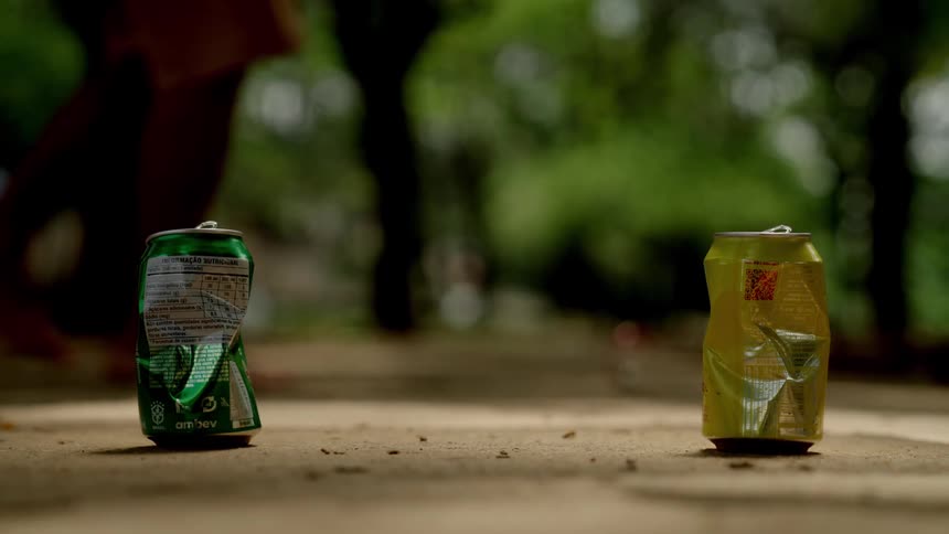 Feet of a person playing with a crushed can on a sidewalk with two cans standing upright