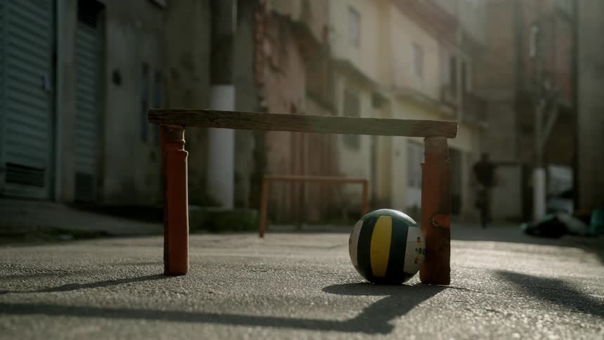 A soccer ball rests beside a makeshift wooden goal in a sundappled alleyway as a person on a bicycle passes by in the background