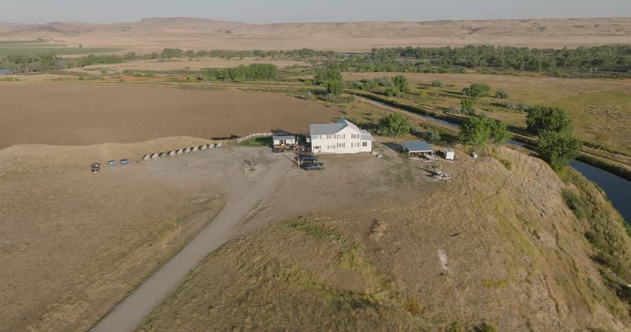 A white farmhouse sits isolated in a dry rural field with trees and a stream nearby