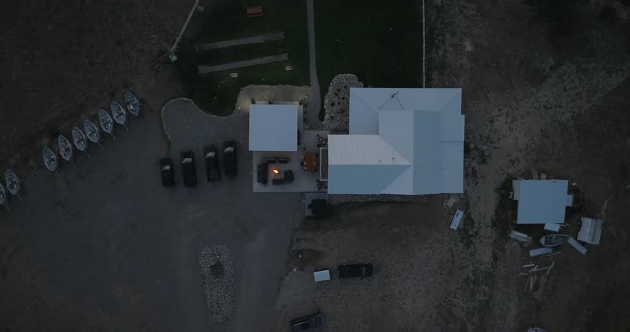 Building complex with white roofs a lit fire pit encircled by couches dirt ground with sparse vegetation parked cars nearby and small boats lined up at the edge under a dim sky