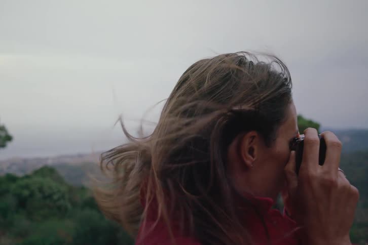 A woman has her hair moving in the wind and she puts a small camera to her face to take pictures