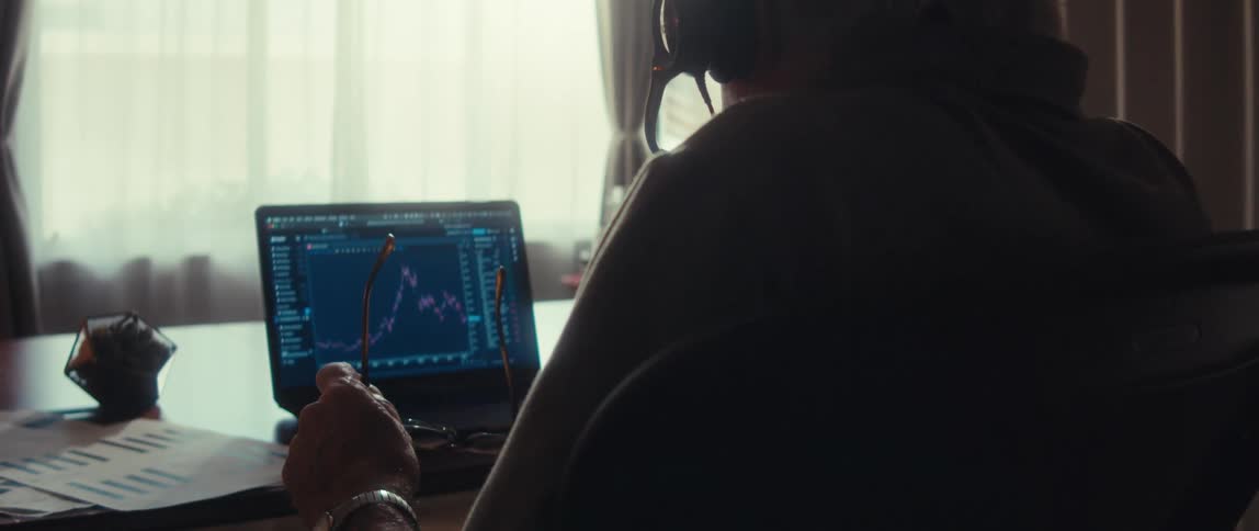 A man sits behind a desk wearing headphones holding his eyeglasses and staring at a computer displaying a stock price chart