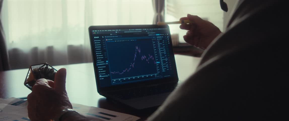Close up of a laptop displaying a trading interface on a table in front of an elderly man demonstrating while speaking