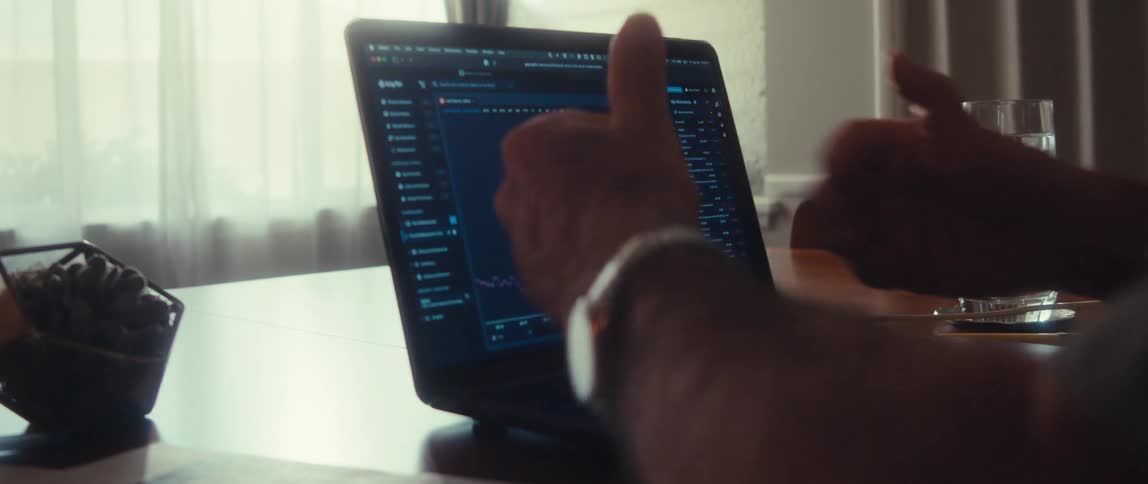 Close up of a laptop on a table displaying a trading software in front of an elderly man speaking and demonstrating with his hands