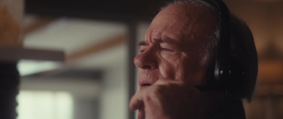An elderly man with glasses and headphones is captured in a close up looking intently and seemingly engaged in a conversation