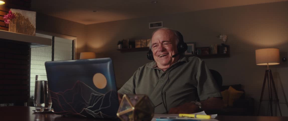 The face of an elderly man who is sitting in a chair looking at a laptop and wearing headphones and laughing and then he takes off his reading glasses