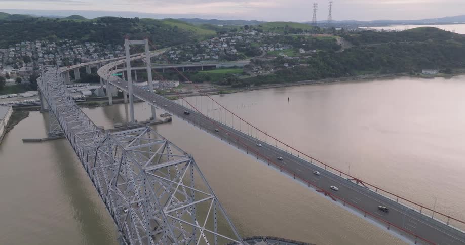 An aerial view shows a long gray bridge with multiple lanes of traffic crossing a wide body of water connecting to a landmass with hills and buildings under a cloudy sky