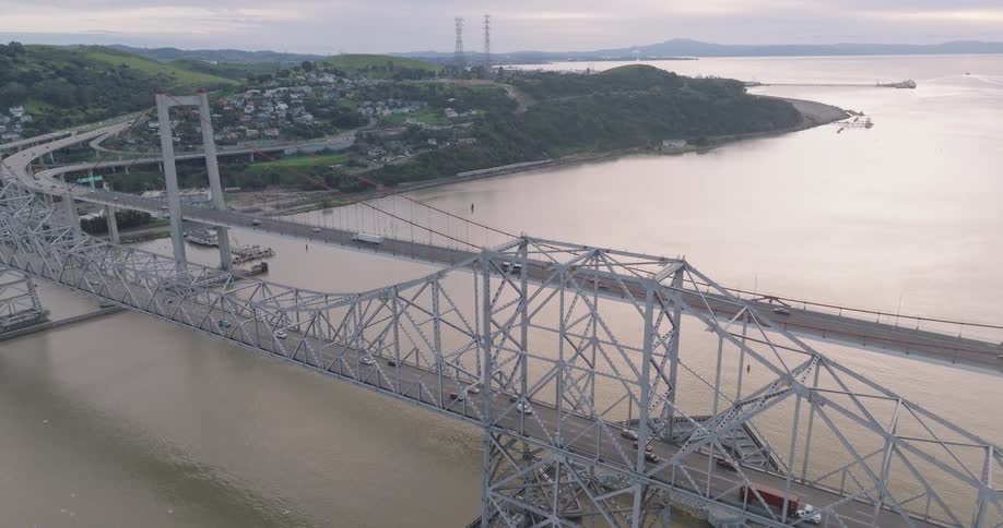A long metal bridge with vehicles crosses over a body of water connecting to land with hills buildings and a distant shoreline
