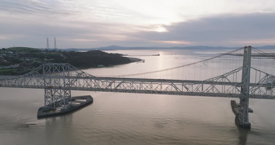 An aerial view showcases a long steel truss bridge spanning a wide body of water under a cloudy sky at dusk with distant hills and islands visible on the horizon