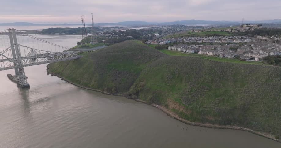 A bridge meets a green hill by the water with suburbs behind