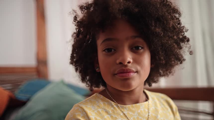 A young girl in a yellow dress looks calmly at the camera in a cozy indoor setting