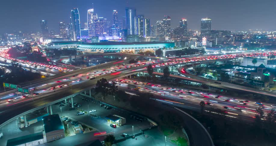 Nighttime traffic flows through a busy Los Angeles highway interchange under a glowing skyline