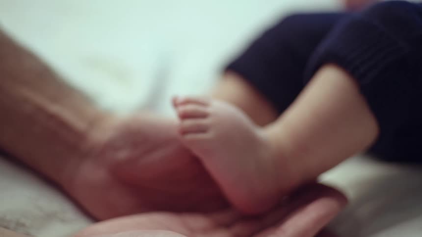 A father holds his babys feet as the child lies on a bed at midday