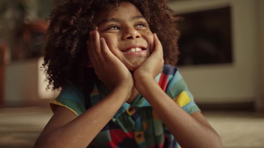 A young child with curly hair resting their chin on their hands and smiling dreamily