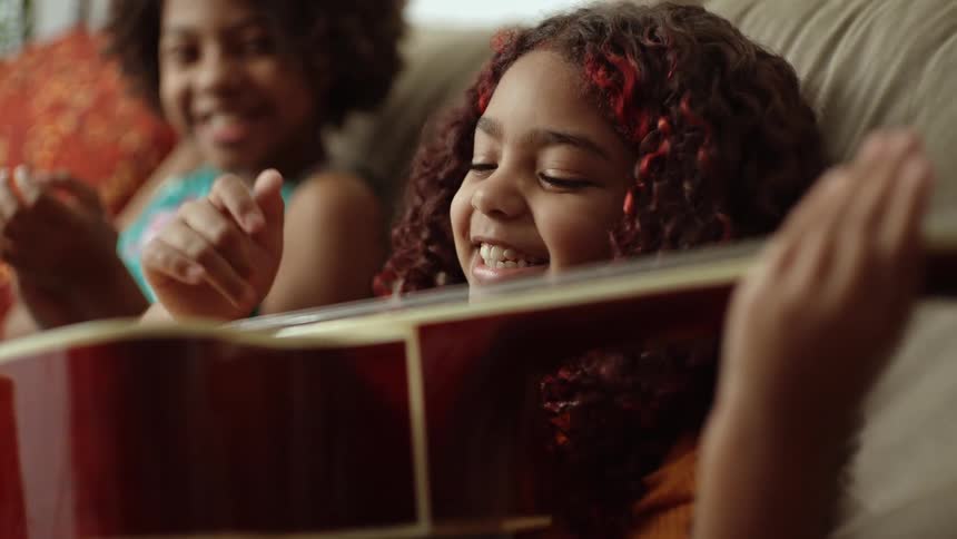 Two young girls joyfully laugh and play guitar on a cozy couch