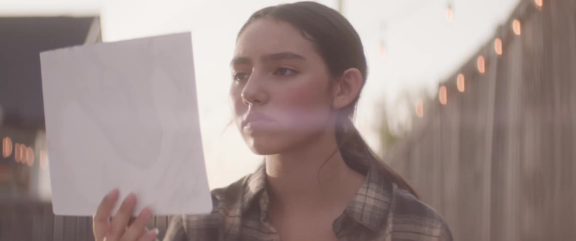The face of a teenage girl who is holding a paper and she is in an outdoor environment with lights in the background