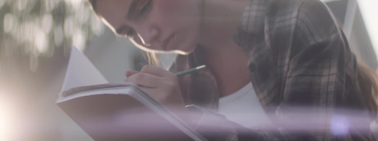 The girl leans close to her notebook drawing with a pencil