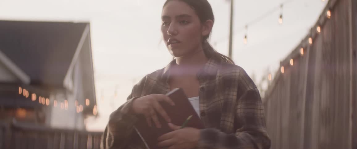 Young woman in a plaid shirt sits in a backyard at sunset finishes writing and holds a notebook tightly to her chest