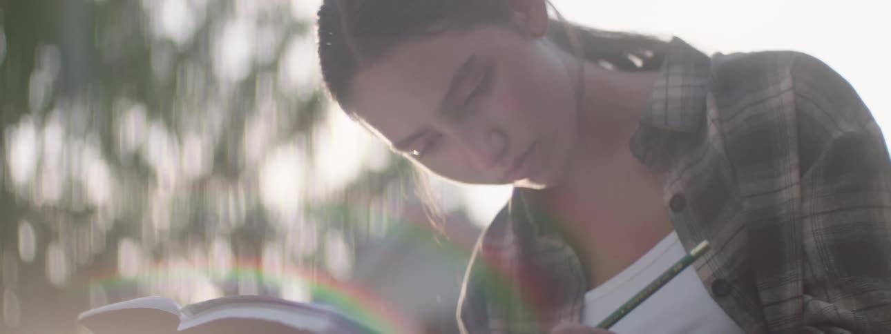 The face of a young woman who is writing in a red cover book in an outdoor setting and she is wearing a plaid shirt
