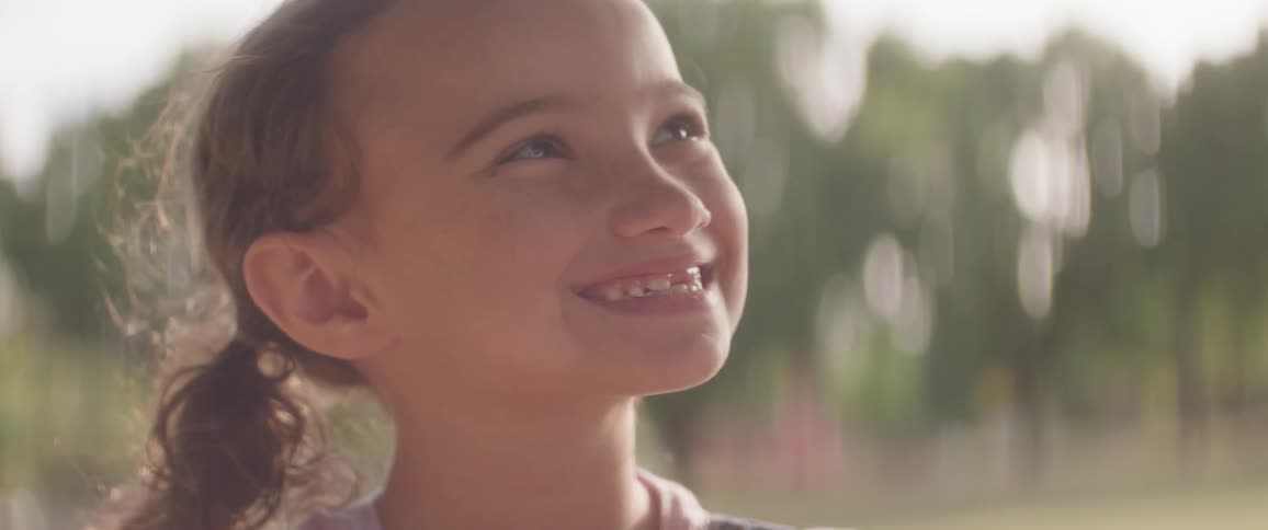 A young girl smiles brightly while looking up in a sunlit outdoor setting