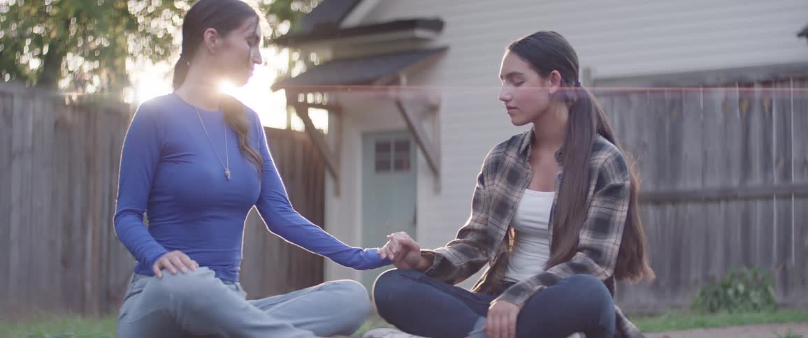 Two women sit cross legged on the grass holding hands in a quiet emotional moment