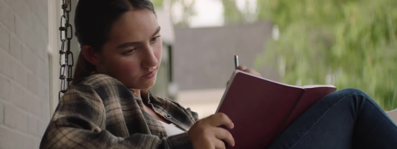 A teenager in a plaid shirt and jeans writes in a notebook while sitting on a porch swing beside a brick wall