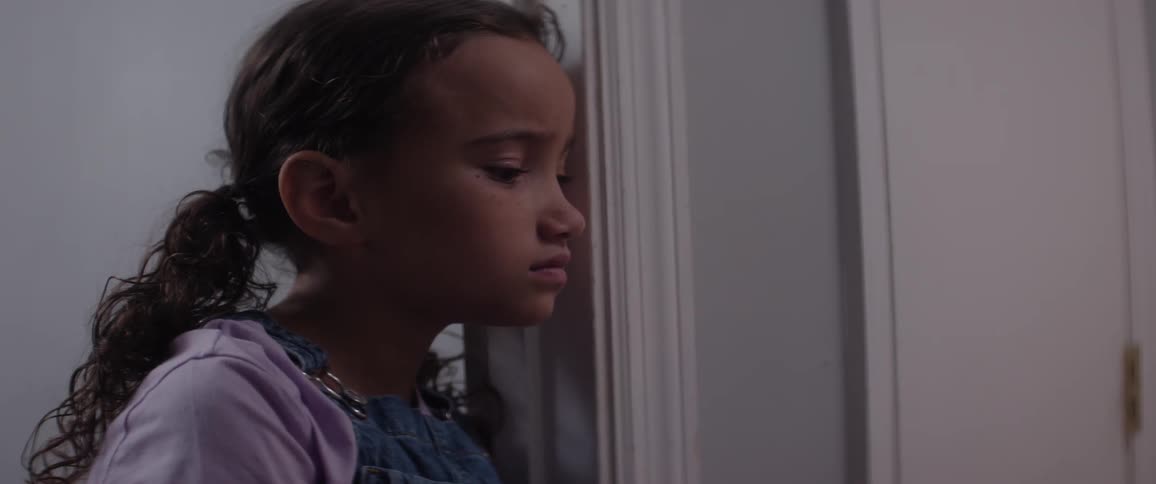 A sad girl with curly hair stands beside a door in a narrow hallway wearing a purple shirt under denim overalls